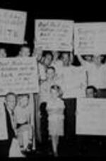 White men and children hold protest signs reading: "Don't Push Our Children Out the Back Door and Let  the Communists Thru the Front Door", "We Belong Advancement of White People", and "Segregation Was Planned Long Before Supreme Court Justices Were Born".
