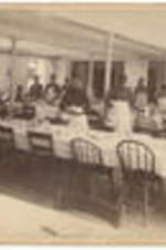 Interior of the dining room in North Hall with tables covered in cloths and stacked with dishes, and women standing throughout the room.