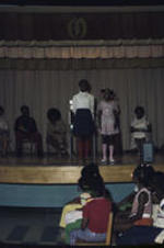 Two unidentified girls stand in front an audience of other children.  A group of unidentified adults sit in a line of folding chairs behind them.