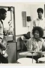 Three young men play musical instruments, bass guitar, drums, and keyboard, in a classroom.