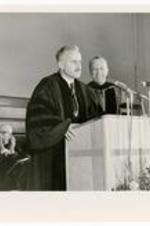 An unidentified man speaks at a podium while Hugh Morris Gloster looks on.
