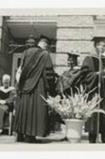 Two men and a woman, wearing graduation caps and gowns, shake hands on an outdoor stage at commencement.