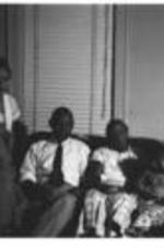 An unidentified group with a baby sit together on a couch in front of a window.