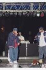 Two men, wearing athletic jackets and baseball caps, hold microphones on stage, a disc jockey and other men stand in the background.