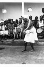 An elderly woman dances topless in ritual while a crowd looks on.