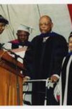 Thomas W. Cole, Jr., Hank Aaron, Xernona Clayton and another man, wearing graduation cap and gowns, stand at the podium at commencement.