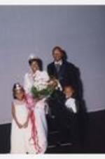 A young woman, wearing a floor-length dress with a tiara and bouquet of flowers, poses with a man and two children on stage.