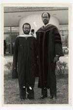 President Elias Blake Jr. and another man, wearing graduation gowns, pose in front of a brick building "Health and Physical Education" at commencement.
