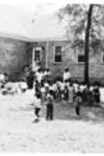 Unidentified children stand outside in the shade under some trees.
