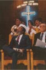 Dick Gregory is shown with Claud Young (at right) and an unidentified man sitting in front of a choir at a Southern Christian Leadership Conference Spring Board meeting held at Grace Temple Baptist Church in Detroit, Michigan.