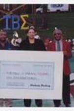 Dr. Walter Boradnax poses with another man and a woman holding a large check "Wachovia Banking, September 28, 2002, Pay to CAU Dept. of Athletics, $5,000," on football field.