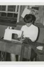 A young woman sits at a Singer sewing machine with a plaid shirt in a classroom.
