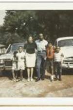 An unidentified, well-dressed family stand in front of two cars. Written on verso: 9-4-67.