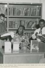 An unidentified group looks at books in a library. Written on recto: An intellectual moment in the library when principals select ideas that may be carried back home, and find that books are important resources.