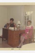 Two women sit at desks by a window.