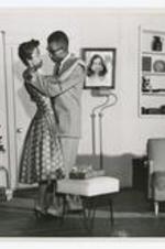 View of man and woman on stage embracing around living room furniture and a small Christmas tree.