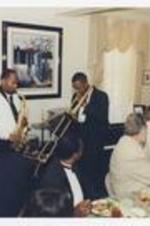 Wynton Marsalis plays his trumpet with a band of four men, playing saxophone, piano, upright bass, and a trombone, in a dining room, while men and women sit at dining tables.