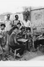 Palestinian refugee children play with bottlecaps as other men look on and smile at the camera.