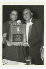 Portrait of man and woman with plaque dedicated to Cleopas Johnson. Written on verso: "A booster member + Mr. Cleopas Johnson- Band Director at Banquet for 20 years of service 1983".