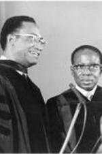 Dr. Reddick and President Senghor stand behind a podium in regalia as C. Eric Lincoln receives his honorary hood in a ceremony at Saint Michael's College..
