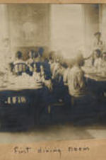 A group of children sit at dining tables. Written on recto: First dining room.