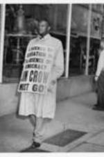 A man walks in front of a store wearing a sign that reads, "The presence of segregation is the absence of democracy, Jim Crow must go!".