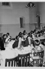 A group talks and eats inside a dining hall. Written on verso: Dining hall event.