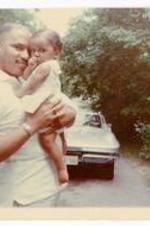 Gladstone "Mickey" Chandler, Jr. holding Beth Angela Warren as a toddler in front of a car. Written on verso: July 4, 1967. Age 1 1/2 mos. Uncle Mickey and I posed in front of Bev &amp; Bill Bart[?]'s car while we were at a picnic at their home. Granma gave me this cute sailor dress. Mickey still "possessive" role.