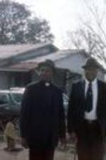 Two male churchgoers, one wearing a priest collar, stand outside with other parishioners. Unknown location.