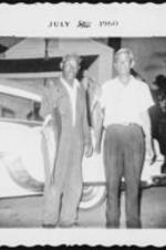 Two unidentified men stand in front of a car and hold fish on lines.