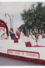 Seven women, wearing ball gown dresses, tiaras and sashes, sit on a red and white parade float with banner "Miss CAU &amp; Court" on street.