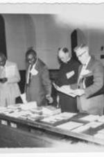 Four unidentified men stand looking at reading material on a table. Written on verso: " Georgia Council of Churches 2/28/58".