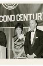 Hugh Morris Gloster stands with his wife, wearing dress with corsage, next to a man delivering a speech. A banner in background reads, "The Second Century"