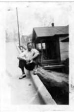 An unidentified man sits on a curb with a row of houses behind him.
