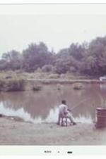 An unidentified man sits on a stool and fishes by a lake.