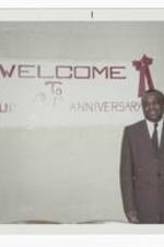 An unidentified man wearing a brown suit stands in front of a white sign reading "Welcome To Our 64th Anniversary."