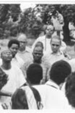 An unidentified man at the center of a circle speaks as others listen.