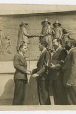 Men shake hands men in front of a stone wall carving.