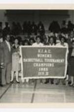 Men and women, including Elias Blake Jr., hold banner "S.I.A.C. Women's Basketball Tournament Champions 1985 Div. III" on basketball court.