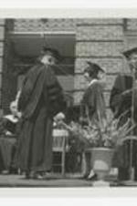 Two men and a woman, wearing graduation caps and gowns, shake hands on an outdoor stage at commencement.