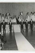 Indoor view of women wearing ball gowns on stage, and men wearing tuxedos standing in front of stage.