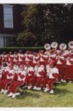 Men and women, wearing red and white marching band uniforms, play musical instruments while sitting and standing in front of folding chairs outdoors at convocation.