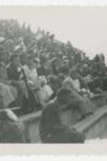 A group sit on the bleachers and watch a football game. Written on verso: Fans at Morehouse College football game.