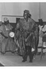A man in African wardrobe dances while men play drums in the background.