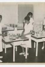 Four women, wearing shirt dresses, stand at two tables with mixing bowls, cutting boards and other kitchen utensils, in a classroom kitchen.