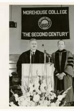 Hugh Morris Gloster stands next to an unidentified man on stage delivering a speech. On banner "Morehouse College, The Second Century".