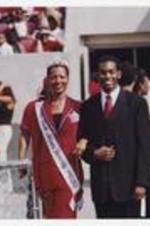 A young woman, wearing a red suit with a tiara and sash "Miss Clark Atlanta University 2002-2003," stands next to a young man, wearing a suit with necktie, in the football stadium, audience members sit and stand in the background.