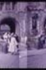 Entrance of a castle with men and women entering the grounds. Two men stand guard next to a car, one wearing a kilt.