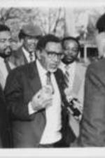 Southern Christian Leadership Conference (SCLC) President Joseph E. Lowery is shown being interviewed by the press at a Winn-Dixie protest. Written on verso: Dr. Lowery explains "evils" of South African canned peaches being sold by Winn-Dixie as ministers look on.