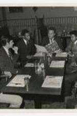 View of six men seated at dining table, view of dining booths in background.